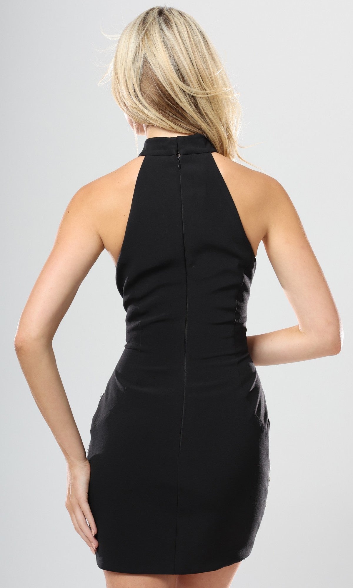 A woman is seen from the back in the Jovani 46402 Short Homecoming Dress—a sleeveless, fitted black semi-formal design—resting one hand on her hip with light-colored hair partially covering her face.